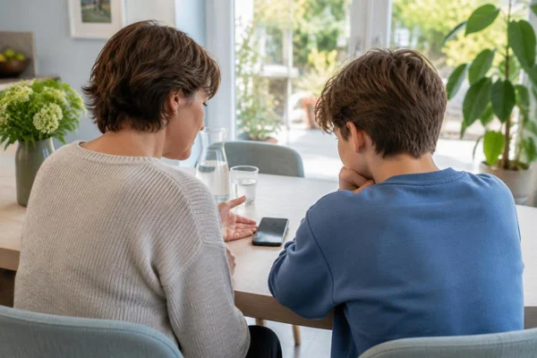 Illustratief beeld: ouder en tiener van achteren aan een eettafel met een smartphone tussen hen
